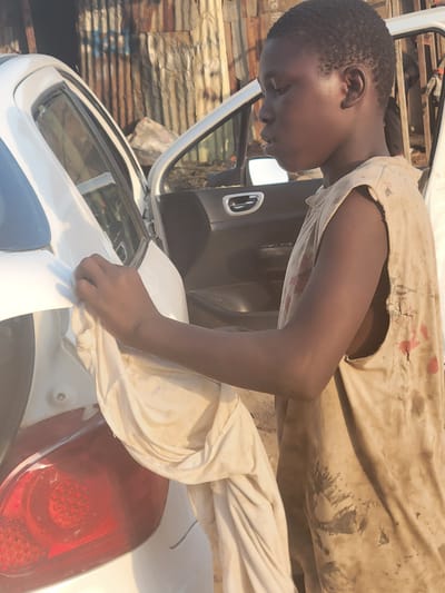 A young, dark-skinned person in Kwamba, Nigeria, is polishing the rear of a white car with a light-colored cloth. The person wears a tattered, stained beige tank top and light-colored pants. The car's open door suggests it might be undergoing cleaning or repair. The background includes corrugated metal sheets, indicating a simple structure, and sunlight is illuminating the scene.