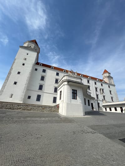 This is a low-angle, vertical photograph of Bratislava Castle in Bratislava, Slovakia. The castle is a large, white building with red-tiled roofs and several prominent towers. The architecture is grand and imposing, with multiple windows and decorative elements. The foreground is paved with cobblestones, and a stone wall runs along the base of the castle. The sky above is a bright blue with scattered white clouds, suggesting it is daytime. The scene is tranquil and hints at a pleasant day for exploring a historical landmark. There are a few people visible on balconies and in the courtyard, casually observing the surroundings. The overall impression is one of historical significance and architectural beauty.