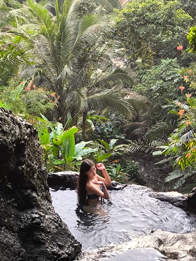 A woman sits in a dark, murky pool of water surrounded by lush, green vegetation, likely in Sidemen, Indonesia. The setting appears to be a natural spring or pool formed among rocks, with tropical plants and trees, including a tall palm tree, densely surrounding it. The woman is wearing a bikini. She has long, dark hair. The lighting is natural. The foreground features dark, rough rocks. The setting is likely a secluded or natural bathing area. A stream is visible behind the pool.