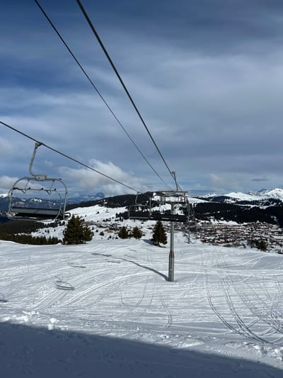 The image depicts a snowy mountain landscape at a ski resort in Villard-sur-Doron, France, under a partially cloudy sky. In the foreground, a snow-covered slope is marked extensively with ski tracks. A ski lift system traverses the scene, with two empty, grey metallic chairs prominently visible; one is in the mid-left foreground, and another is further right, positioned near a grey metal support pylon that casts a shadow onto the snow. The ski lift cables extend diagonally across the upper portion of the frame. In the midground, evergreen trees are scattered across the snow-covered terrain. Below and beyond the ski lift, a village consisting of numerous buildings with brown roofs is visible, nestled in a valley. The background features additional snow-covered mountains and forested hills under a blue and white sky. No individuals are present in the scene.