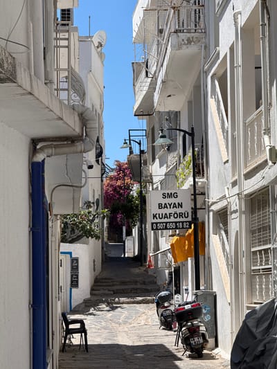 This is a vertical, eye-level shot of a narrow, sun-drenched alleyway in Bodrum, Turkey. The scene is dominated by whitewashed buildings with blue accents, typical of the region's architecture. Several objects are visible, including two scooters parked on the right side of the alley, a black fabric cover draped over something in the upper right, and various pipes and conduits running along the exteriors of the buildings. A sign for "SMG BAYAN KUAFÖRÜ" (SMG Ladies' Hairdresser) with a phone number is hanging from a black pole. Yellow fabric is draped over a clothesline above the sign. There are also glimpses of green foliage and pink flowers adding a touch of color to the otherwise monochromatic buildings. The sky is a clear, bright blue, suggesting a sunny day. The perspective is from a low angle, looking up the alley, which recedes into the distance. The image conveys a sense of everyday life in a Mediterranean town. There are license plates visible on the scooters with text like "48 VT 835" and "48 8191".