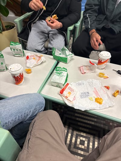 A table covered in fast food wrappers and containers suggests a recent meal. Used cups, crumpled paper bags labeled "Onion Rings," and discarded food containers litter the surface. Two people are seated at the table, one is eating fries and the other is wearing a dark jacket. Another person with blue jeans is seated at the table as well. A patterned floor is visible.