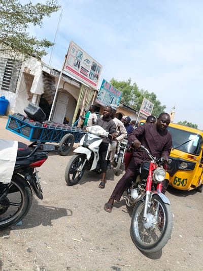 The image depicts a street scene in Gama, Nigeria, under a bright, clear sky. Multiple individuals, predominantly adult males, are present on a dusty or unpaved road. In the immediate foreground, a man wearing a dark brown long-sleeved garment and dark pants sits on a red and chrome motorcycle, looking towards the camera. He wears dark flip-flops. Directly behind him, another man in a dark grey shirt and dark pants is seated on a white and black motorcycle, also facing forward. Several other men on motorcycles are visible further back in the line of vehicles. To the right, a yellow auto-rickshaw (Keke Napep) with the number "6543" on its side is partially visible. To the left, a blue metal two-wheeled trailer, laden with multiple black jerrycans or containers, is parked or being towed. One black container rests precariously on top of the stack.
The background features a dilapidated building with exposed brickwork and a corrugated metal roof on the left, near which a blue barrel or bin is visible. Above this building, a large white billboard sign, advertising what appears to be various products or services, is mounted. A smaller blue sign is positioned below it. Further back, green trees are visible, and in the far background on the right, a light-colored mosque minaret with a pointed top rises against the sky. A tall, slender antenna or mast is visible in the distance on the far left. The scene suggests a busy thoroughfare or commercial area with pedestrian and vehicular traffic.