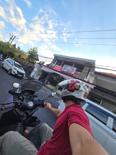 The image depicts a person riding a motorcycle in Tibubeneng, Indonesia. The rider is wearing a helmet and a red shirt, and is seen from a first-person perspective, looking down the road. The motorcycle has a black windshield and mirrors. A white car is parked on the side of the road, near a building with a store that appears to sell bottled water and beverages. Overhead power lines stretch across the sky, which is blue with scattered clouds.