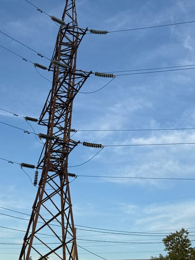 This media file shows a low-angle, wide shot of a high-voltage power line tower against a bright blue sky with wispy white clouds. The metal lattice structure of the tower is rusty brown and takes up the lower portion of the frame, stretching diagonally from the bottom left to the top right. Several thick, black electrical wires are suspended from the tower, extending upwards and out of the frame. Ceramic insulators, ribbed and translucent, are visible along the tower, attached to the wires.
In the upper right corner of the image, a small section of green foliage from a tree is visible, suggesting an outdoor setting. The lighting indicates it is daytime, likely late morning or early afternoon, given the clear sky and bright illumination. There is no visible text in the image. The scene evokes a sense of industrial infrastructure against a natural backdrop. The focus is on the massive scale of the power lines and the tower, with the sky providing a vast expanse. No people or specific activities are depicted. The location context of Petropavl, Kazakhstan, hints at a potentially rural or semi-urban setting where such infrastructure would be common.