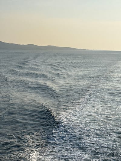 The image displays a maritime scene in the vicinity of Palaiopoli, Greece. The upper approximately one-fifth of the frame presents a hazy, pale yellow-beige sky. Below this, a dark, indistinct landmass, characterized by rolling hills or mountains, extends across the distant horizon. The predominant feature is the expansive body of dark blue-grey seawater, occupying the majority of the image. The water's surface exhibits a pattern of parallel ripples and waves. A distinct, diagonal trail of white, frothy wake, generated by an unseen vessel, originates from the lower right quadrant and extends towards the upper left. This wake visibly disrupts the surrounding wave patterns. No individuals, specific structures, or identifiable vessels are present within the frame.