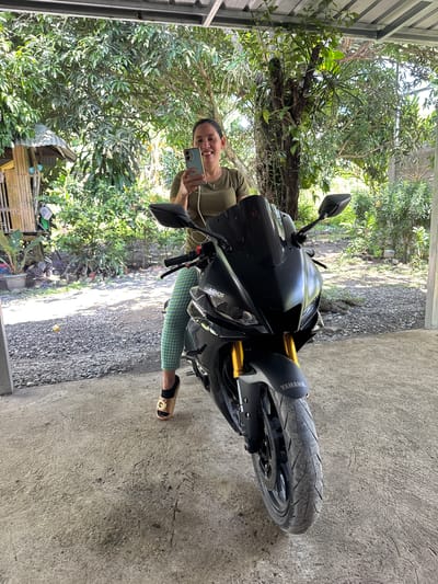 An adult female is seated on a matte black Yamaha R3 motorcycle, positioned on a concrete slab in a semi-covered outdoor area in Carmen, Philippines. She is smiling and looking towards the viewer, holding a light blue/green smartphone with both hands, appearing to engage with its screen. She wears an olive-green short-sleeved top, green and white checkered capri pants, and a beige sandal with a black strap and gold-colored buckle on her left foot. The motorcycle, adorned with "YAMAHA" on the front fender and "LUMINOS" on the side fairing, features gold-colored front forks and is facing the viewer. The immediate ground under the motorcycle is concrete, transitioning to a gravel and dirt path beyond. To the left background, a rustic bamboo structure with a corrugated metal roof is visible, accompanied by potted plants. The background is dominated by dense, lush green tropical vegetation, including large trees and various bushes, indicative of a garden or rural setting. Overhead, a metal roof structure provides partial cover. The scene is illuminated by natural daylight.