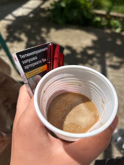 A high-angle shot depicts a hand holding a white, ribbed, plastic cup filled with a light brown liquid, presumably coffee or tea with milk. A pack of cigarettes rests against the cup, with the warning "Тютюнопушенето запушва артериите Ви" (Smoking clogs your arteries) visible, along with a hotline number for quitting smoking. The background is blurry, but suggests an outdoor setting, possibly a yard or sidewalk. Based on the text on the cigarette pack, the image is likely taken in Bulgaria, specifically Koniovets, Bulgaria.