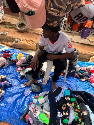 In Nyanya, Nigeria, a street vendor sits on a white plastic chair next to a collection of items for sale, spread across a large blue tarp on the ground. The vendor, a man with dark skin wearing a white t-shirt with red trim and black pants, is partially obscured by the items. He is seated and appears to be looking away from the camera.
The items for sale include numerous caps and socks. Several colorful caps are piled on the left side of the tarp, with some featuring animal ears and colorful designs. A variety of socks, many with playful and patterned designs, are spread out across the rest of the tarp. Some socks display cartoon characters, animals, and holiday-themed patterns. There are also black plastic bags, a beige sack filled with goods, and a used tire visible in the background. The ground surrounding the tarp is uneven and sandy, typical of an outdoor market environment. In the distance, a sign with text and images is visible, and what appears to be a small propane gas cylinder is also present. The overall scene depicts a busy street market atmosphere.