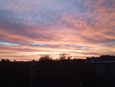 This media file depicts a picturesque sunset scene over a residential area in Kyshtym, Russia. The sky is a canvas of vibrant colors, with streaks of pink, orange, and blue clouds stretching across the frame. The clouds are wispy and layered, suggesting a dynamic and ever-changing atmosphere. In the distance, silhouetted buildings and trees create a dark, contrasting foreground against the colorful sky. The architecture appears to be typical of a Soviet-era apartment complex, with flat roofs and numerous windows. The presence of antennas on the rooftops further reinforces this urban setting. The overall mood is serene and tranquil, evoking a sense of peace and natural beauty. The time of day is clearly evening, as indicated by the sunset and the dim lighting on the buildings. The weather appears to be clear, allowing for an unobstructed view of the sky's spectacle. There is no visible text in the image.