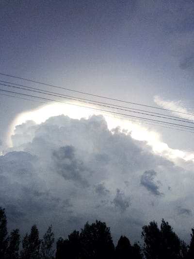 The image depicts an overcast sky over Nachu ward, Kenya, dominated by a large, dense cloud formation. The upper portion of the sky is a muted bluish-grey, transitioning to a brighter, whitish-grey hue towards the horizon line where a significant light source, likely the sun, is obscured by or piercing through the clouds. This light source creates a brilliant white outline along the upper and upper-right edges of the central, dark grey and bluish-grey cumuliform cloud. Four distinct, dark utility lines traverse horizontally across the upper-middle section of the frame, positioned above the brightly illuminated cloud edge. The lower quarter of the image features the dark, silhouetted canopy of numerous trees, appearing as an indistinct jagged mass against the illuminated clouds. No individuals, animals, or specific ground-level structures are visible. The overall scene conveys dramatic natural lighting conditions with visible image grain.