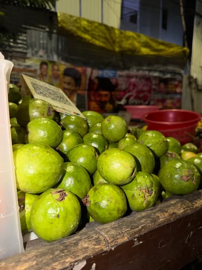 A close-up, angled shot focuses on a pile of green guavas. The guavas are round and have a slightly textured skin, with some showing small brown blemishes. They are densely packed in a white plastic container, with a rough, dark wooden beam to the right, suggesting a market stall or fruit stand. In the background, a red bucket is visible, and behind that, posters with images of people are displayed, hinting at a commercial setting. A QR code on a white card is partially visible in the foreground, to the left of the guavas. The lighting suggests it might be evening or night, with artificial light illuminating the scene. The overall impression is a vibrant display of fresh fruit in a busy, possibly outdoor, market environment in Chennai, India.