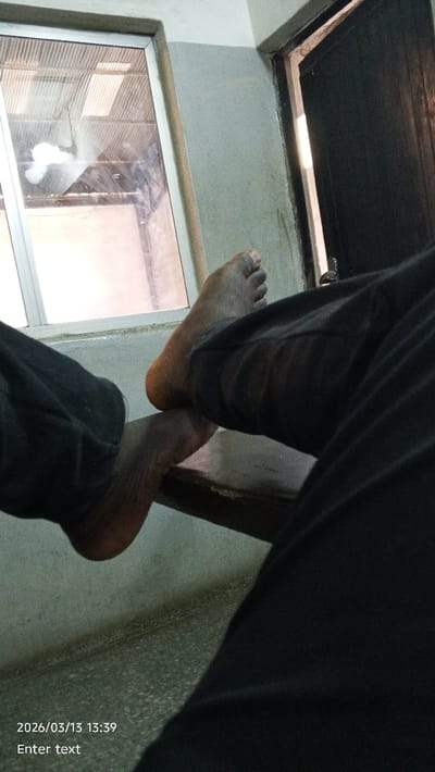 The image displays an indoor scene in the city of Dakata, Nigeria, observed from a low, seated vantage point. In the foreground, the lower legs of an individual are visible, clad in dark trousers. The person's bare, dark-skinned left foot rests on top of their bare right foot, which is positioned on a dark, flat surface, likely a bench or table. The background features a light grey wall, bisected by a darker grey horizontal band near the top. To the left, an aluminum-framed window reflects an interior corrugated ceiling structure and shows an obscured view of an exterior environment. To the right, a dark-colored door is partially open, revealing a vertical strip of bright light from an adjacent area and a metallic door latch mechanism. The floor, visible in the lower left, is light-colored and exhibits a textured surface. A digital timestamp "2026/03/13 13:39" and the text "Enter text" are superimposed on the bottom left corner of the image.