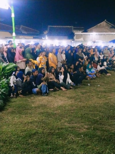 A group of people are gathered on a grassy field at night, in Kecamatan Sambirejo, Indonesia. The field is illuminated by overhead lights and there are buildings in the background. The group is comprised of approximately 50 people, most of whom are seated on the grass. Some people are standing and chatting with others. They are all facing forward and there are no visible interactions or actions taking place between them. There are numerous plants in the foreground, and the group is seated in front of them. The people in the group appear to be of various ages and wearing a variety of clothes. The majority are wearing traditional clothing and there is a mix of men and women. The environment is dark and the atmosphere is relaxed.