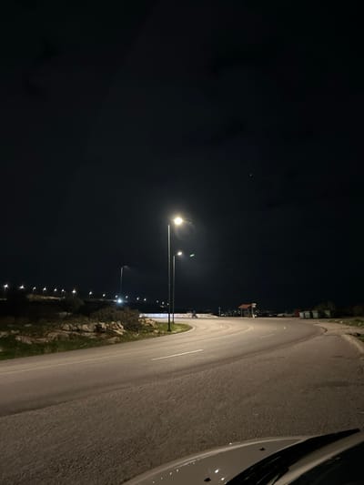 The image displays a nocturnal scene of a curving road illuminated by artificial light, set against a dark sky. The perspective appears to be from a low angle, possibly from a stationary vehicle or a person standing next to one, as a small section of a white car is visible along the far right edge of the frame.

A paved asphalt road, featuring a white dashed line, curves from the foreground towards the middle distance on the right side of the image. Alongside the road, on the right, there is an embankment characterized by rocks and sparse green vegetation, partially highlighted by the streetlights. Several streetlights are present, with two prominent poles standing in the mid-ground on the left side of the road, casting bright, warm-toned illumination onto the road surface and its immediate surroundings. Further into the background, a succession of smaller lights extends into the distance, suggesting a distant town, city, or a continuous stretch of road.

The sky, occupying the upper left quadrant, is very dark and appears mostly clear, with a single faint light source, possibly a star or a distant airplane, visible. The overall atmosphere conveys quiet solitude, characteristic of a road late at night. There are no people, animals, or other vehicles immediately visible in the scene. The weather seems clear, with no indications of fog, rain, or significant cloud cover. This scene is located in Anatoliki Attiki, Greece. No visible text is present within the image.