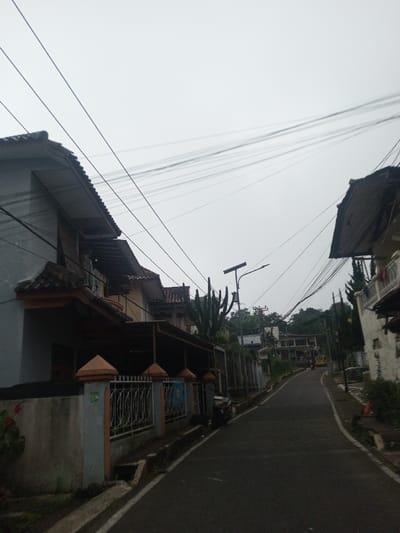 A street scene shows a narrow asphalt road curving uphill, lined with houses on both sides. The houses are various styles, some with covered porches and decorative fences. Power lines crisscross overhead against a gray, overcast sky. Vegetation includes trees and a tall cactus. A small yellow vehicle is visible further up the street. The image suggests a residential area, possibly in a hilly or mountainous region.