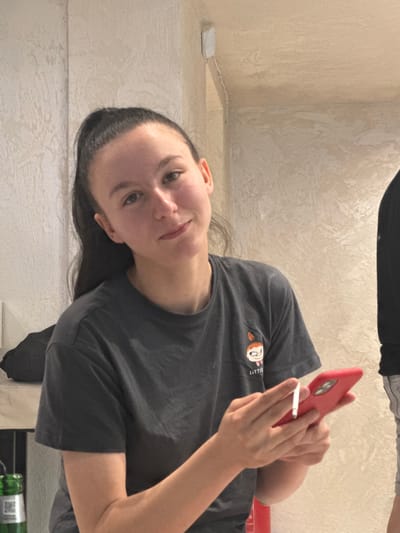 In a room with textured, light-colored walls, a young woman with dark hair pulled into a high ponytail is holding a red smartphone and a lit cigarette. She is wearing a dark gray t-shirt with a small graphic of a cartoon character on the left chest. The woman is looking towards the camera with a slight smile. To her right, another person, mostly out of frame, is visible wearing a black t-shirt and light-colored shorts. In the background, to the left, a green bottle is partially visible. The overall impression is an indoor, casual setting. No specific location in Lom, Bulgaria can be determined from the image.