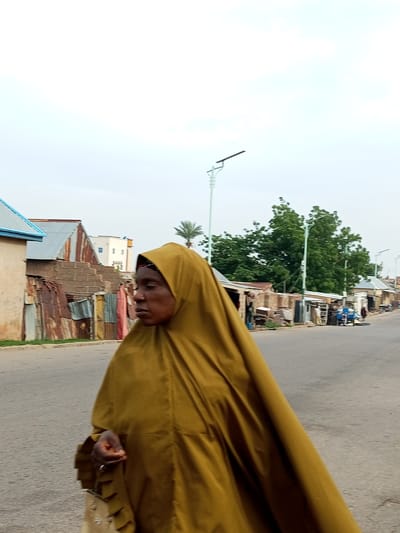 In Arawa, Nigeria, a woman wearing a mustard-yellow hijab walks on a paved road. She is walking from left to right, and her head is turned to her left, looking away from the camera. She is carrying a light-colored bag, possibly a purse, with a strap that rests on her left shoulder. Her right hand is visible, holding the bag strap. The background features a street lined with buildings and trees. Some of the buildings appear to be constructed with corrugated metal and brick. There are streetlights visible, and a few people and a cart are in the distance. The sky is overcast.