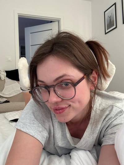 The media file shows a close-up, tilted selfie of a young woman lying in bed. She has brown hair pulled back in a messy ponytail and is wearing glasses with dark frames. Her head is tilted and she is looking towards the camera with a slight smile and her mouth slightly open. She is wearing a grey t-shirt and white socks. The background appears to be a bedroom, with white bedding and a hint of a doorway and a framed picture on the wall. The lighting suggests it could be daytime. The scene appears to be a candid, casual moment.