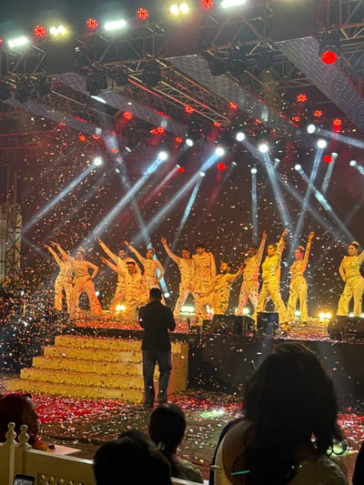 A stage show unfolds with performers in white outfits striking poses amidst a shower of confetti. Bright stage lights illuminate the scene, creating dramatic spotlights and beams that pierce through the air. A metal structure stands on the left side of the stage, adding an industrial element. The audience is partially visible in the foreground. A person in dark clothing stands between the audience and the stage, presumably taking a photo.