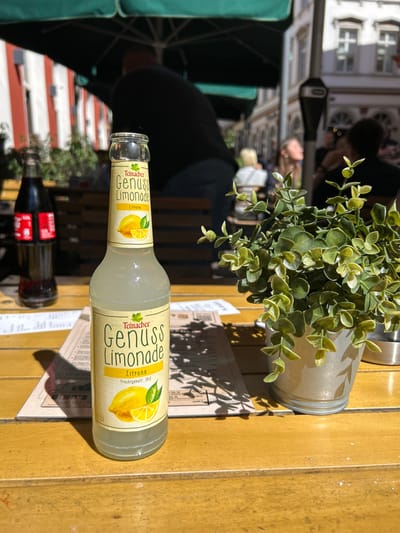 Here's a detailed description of the image:

**Setting:** The image appears to be taken at an outdoor cafe in the city of Heidelberg, Germany. This is suggested by the architecture visible in the background and the style of the cafe setting.

**Objects:**
*   A bottle of "Teinacher Genuss Limonade Zitrone" (lemon-flavored lemonade) lies horizontally on the table.
*   A potted plant is also placed on the table.
*   A restaurant menu sits underneath the lemonade bottle.
*   A dark bottle is seen beneath the lemonade bottle.
*   The table is made of wood.

**People:**
*   The presence of people is implied through the setting (a cafe) and some blurry figures in the background. However, no faces are clearly visible.

**Environment:**
*   The environment appears to be a sunny day at a typical cafe.
*   The architectural style suggests a historic European city like Heidelberg.
*   The presence of an umbrella implies an outdoor cafe seating area.
