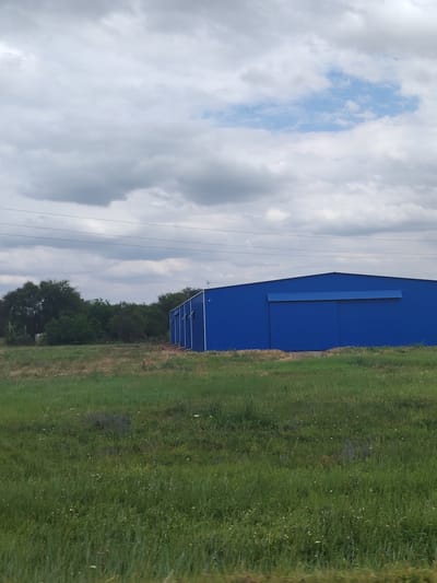 A blue industrial building is located in an open, grassy field under a cloudy sky. The building is rectangular with a sloped roof and appears to be a warehouse or storage facility. There are several windows along the side of the building. In the background, there is a line of trees and power lines. The field is covered in green grass with some scattered wildflowers. The overall scene suggests a rural or semi-rural industrial area.