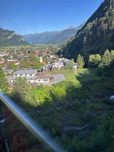 This is a high-angle, wide-shot, landscape photograph of the village of Hüsen, Switzerland, taken on a sunny day. The foreground is dominated by a lush, green, forested mountainside and a small stream. A rainbow-like glare streaks across the lower left portion of the image. In the midground, several buildings are visible, including a light-colored, rectangular building with a silver roof covered in solar panels. A church with a dark steeple is visible in the distance. The background features a wide valley with a small town nestled within it, surrounded by green fields and imposing, rocky mountains. The sky is clear and blue. No people are visible in the photograph.