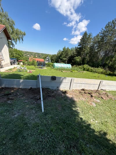 The image depicts a residential backyard in Pisanets, Bulgaria, under a clear blue sky with scattered white clouds. The foreground shows a newly constructed concrete retaining wall. To the left, part of a house with a red-tiled roof is visible, along with a tall tree. The middle ground of the yard is covered in green grass and features a trampoline, a small greenhouse, and gardening beds. In the background, a dense line of trees forms the boundary of the property. The overall scene suggests ongoing yard work or construction.