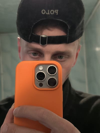 The image, captured in Лабжанскі сельскі Савет, Belarus, presents a close-up mirror selfie of a male subject from the forehead to the upper chest. The subject, possessing light skin and light blue-grey eyes, wears a black baseball cap backward, displaying the inverted and reversed embroidered text "POLO" on the rear, along with a visible metallic strap buckle. A dark-colored garment is worn on the upper body. The subject's right hand holds an orange-cased smartphone, likely an iPhone Pro model, positioned directly in front of the lower half of his face, thereby obscuring the nose, mouth, and chin. The smartphone exhibits a prominent triple-lens camera array and a flash module. The background consists of light blue-green tiled walls with visible grout lines, indicating an indoor setting.