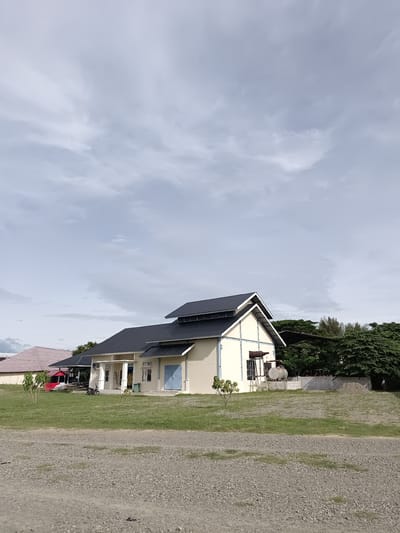 The image depicts a scene in Mesjid Reuntoh, Indonesia, featuring a light-colored, modern building with a dark pitched roof. The building has several windows and a blue door, with an entrance area supported by white pillars. To the left of this main building, another structure with a reddish-brown tiled roof is partially visible. A red, open-sided shelter is situated between the two buildings, with some construction materials and a motorcycle parked nearby. The foreground is a gravel-covered area, transitioning to a grassy lawn with a few small trees. In the background, dense green trees form a natural backdrop. The sky is overcast with scattered clouds. There are no people clearly visible in the image. The overall impression is that of an institutional or commercial property in a semi-rural or developing area.