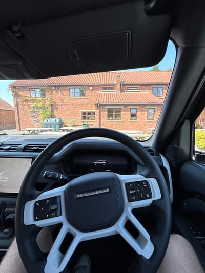 The image depicts a static scene observed from the driver's perspective within a Land Rover Defender, looking towards a residential or commercial building in Wold Newton, United Kingdom. The foreground prominently features the vehicle's interior, including the black and white steering wheel with "DEFENDER" branding and various control buttons. Behind the steering wheel, the digital instrument cluster displays a stylized silhouette of a Land Rover Defender. To the left, a portion of the vehicle's infotainment screen is visible. Below the steering wheel, the driver's lower bare legs and a dark shoe are discernible. An overhead black sun visor is lowered in the upper center of the frame.
Through the windshield, a substantial two-story building constructed of red brick with a red tile roof is visible under a clear, bright blue sky. The building features multiple brown-framed windows of varying sizes across its facade. On the left side of the building, green climbing vegetation grows on a wooden trellis structure. In the paved area directly in front of the building, a large dark green, possibly plastic, outdoor storage container or compost bin is present, with a wooden picnic table and benches situated behind it.