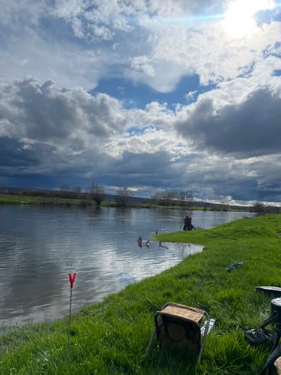 This image captures a wide-angle view of a river landscape in Glava, Bulgaria, under a dynamic sky. The upper portion of the frame features a blend of dark grey cumulus clouds, particularly dense in the upper left, transitioning to lighter, white clouds and patches of clear blue sky towards the upper right, where a bright sun is visible, creating a lens flare.
A wide, dark grey-blue river occupies the mid-ground, flowing from left to right. Its surface reflects the sky, showing ripples and indicating a calm current. The near bank, in the foreground, is covered with lush, vibrant green grass, sloping gently towards the water. Planted vertically in this grass, close to the water's edge, is a red Y-shaped fishing rod rest or holder. Further back from the river on the grassy bank, a camouflage-patterned foldable stool or chair is present, accompanied by several dark-colored bags or fishing equipment items resting on the grass. A clear plastic bottle is also visible on the grass.
The far bank of the river is also green, featuring scattered deciduous trees and bushes, many appearing bare or with minimal foliage. In the distant background, low hills or mountains are faintly visible beneath the darker cloud cover. No individuals are present in the frame. The scene suggests a daytime setting, possibly early spring, with equipment implying recreational fishing activity.