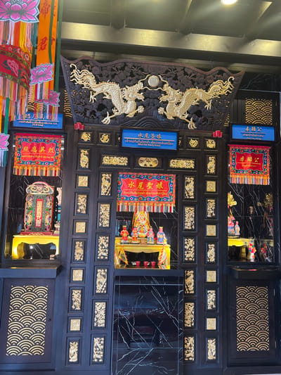 This is a medium-shot, eye-level photograph of an ornate altar within the Wat Hua Thanon in Baan Hua Thanon, Thailand. The altar is situated within a dark, intricately carved wooden structure adorned with gold accents. The structure features two large, golden dragons prominently displayed above the central altar area.
The central altar itself is adorned with several small, colorful statues of deities and offerings. A red banner with gold lettering hangs above the statues. To the left and right of the central altar are two smaller alcoves, each containing a shrine with a statue and offerings, also decorated with red and gold banners. The back wall of the altar area is a dark, reflective material, possibly marble or polished wood, with white veins. The overall impression is one of rich cultural and religious significance.