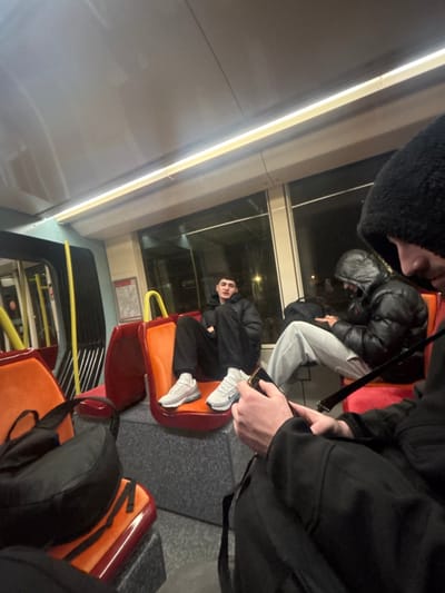 Inside a train carriage, passengers occupy orange and red seats. A young man in a black jacket and black trousers sits with his feet up, wearing white sneakers. Another person, also in a dark jacket, sits behind him in grey sweatpants. A third person wearing a black beanie leans into the frame, partially obscuring the view. The window reveals a night scene with distant lights.