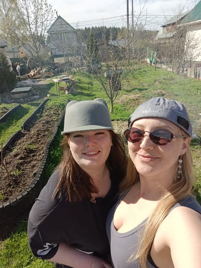 Two women pose for a selfie outdoors in Вичелово, Russia. The woman on the left has brown hair and a gray hat with small ears. She wears a black shirt. The woman on the right has blonde hair, sunglasses, a gray cap with white lettering, and dangling earrings. She wears a gray tank top. Behind them are garden beds. In the background are houses, trees, and a person working near a pile of wood and smoke. The weather appears sunny.