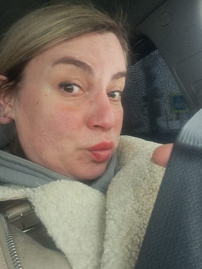 A woman's face fills the frame, captured from a slightly angled, close-up perspective. She has fair skin with some redness on her cheeks, brown eyes with visible eyelashes, and pursed lips with lipstick. Her blonde hair is visible on the side. She's wearing a light grey top and a cream-colored, plush jacket, partly obscured by what appears to be a seatbelt. The background suggests the interior of a car, including a ceiling and sun visor.