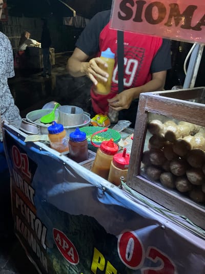 This image depicts a night scene at a street food stall in Kauman, Indonesia. A male vendor, wearing a red and black t-shirt with a "29" graphic, is visible from the chest down, holding an amber-colored squeeze bottle with a light blue cap, suggesting he is dispensing sauce or seasoning. His hands are visible, with dark skin. To his left, a portion of a customer wearing a patterned shirt is visible.

The food cart features a glass-fronted wooden display case on the right, filled with numerous light brown, spherical meatballs (likely pentol or bakso). On the cart's counter, several squeeze bottles with red and blue caps hold various sauces and condiments. Adjacent to these are metal containers, including a pot and a cylindrical vessel, along with a bright green plastic ladle and a green plastic basket containing chopped green vegetables. A translucent plastic bag with contents is also visible on the counter.

Above the vendor's head, a clear sign with red lettering reads "SIOMA," likely referring to "Siomay," an Indonesian dish. The lower part of the cart is covered by a banner featuring text such as "URAT PENTOL KUWAH" and "APA" over a landscape image, along with red circular graphics. The background is dark, indicating nighttime, with faint artificial lighting illuminating two indistinct figures further back, one appearing to be tending to another area. The ground in the background appears wet or reflective.