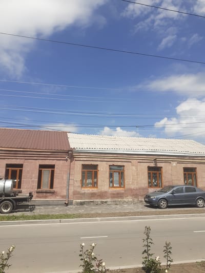 The image shows a roadside view of two attached buildings with different roofing materials. The building on the left has a reddish-brown, tiled roof, while the building on the right has a white, corrugated roof. Both buildings are constructed of stone and have several windows with wooden frames. A dark-colored car is parked in front of the right building, and a water truck is parked in front of the left building. The buildings are situated alongside a paved road with a sidewalk separating it from the buildings. In the foreground, there are some flowering plants. The sky above is blue with scattered clouds, and power lines are visible against the sky. The scene appears to be in a semi-urban or rural area, potentially along the border of Gyumri-Akhuryan, Armenia, given the architectural style and the presence of infrastructure like power lines and a paved road. There are no people visible in the image.