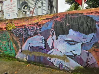The image captures a vibrant mural on a retaining wall, with the "Alianza Francesa Osorno" building visible above it in Osorno, Chile. The mural depicts two contrasting scenes. On the left side, there is a rural scene featuring a group of animals, possibly oxen or cattle, being led along a path. A person can be seen walking among them. Behind the animals is a painting of a yellow building. To the right, the mural transitions into a close-up portrait of a person, rendered in shades of blue and purple. The person appears to be engaged in some form of manual work, with their hands and part of their face visible, and the detail of the person's face is well-executed. The mural is executed in a bold, expressive style, with strong colors and visible brushstrokes. Moss and other vegetation can be seen growing at the base of the wall, near the sidewalk, adding a touch of nature to the urban setting. Additionally, there is a sign visible behind the mural, in white with red writing, detailing information about learning French and cultural workshops.