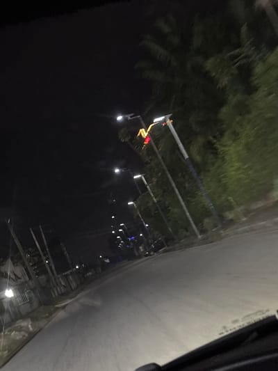 The image shows a dark street at night in Port Harcourt, Nigeria, illuminated by streetlights. There are trees and foliage visible on the left side of the frame. Several streetlights are present, suspended by metal poles. A section of what appears to be a concrete structure or building is visible on the right side of the image. There are no people visible.