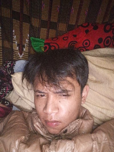 A person lies on a tan pillow, wearing a light brown, shiny jacket. A red pillow with black circles is behind them. Above that is a patterned brown and white tapestry. The person has short, dark hair and looks directly at the camera with a slightly furrowed brow. Their expression is serious. The lighting is dim.