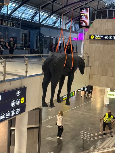The image captures a scene inside a modern airport terminal, likely in Oslo, Norway, given the visible text. The perspective is from a high angle, looking down at the floor and some of the airport's architecture.

The most prominent object is a large, black, abstract sculpture resembling a horse with elongated legs, suspended by orange cables from above. It appears to be part of an art installation or decoration within the terminal.

Several people are visible in the scene. In the upper right, a person wearing a high-visibility yellow vest, likely airport security or staff, is leaning on a railing. Further down, a woman with blonde hair is walking away from the camera. In the background, other individuals can be seen walking or standing. On the left side, near a screen displaying a close-up of eyes with the text "SER DU DETTE?" (Are you seeing this?), people are gathered or passing by. Another group is visible near a "BAGUETTE" and "Coffee" sign in the lower left.

The setting is a spacious, well-lit airport interior with a tiled floor. Signage for "Toilets" with an arrow and icons for different modes of transport like airplanes, buses, and trains are visible, indicating a transportation hub. The overall impression is that of a busy, functional public space with an artistic element. The lighting suggests it could be daytime, but the specific time of day isn't clear. There are no obvious indicators of weather conditions.