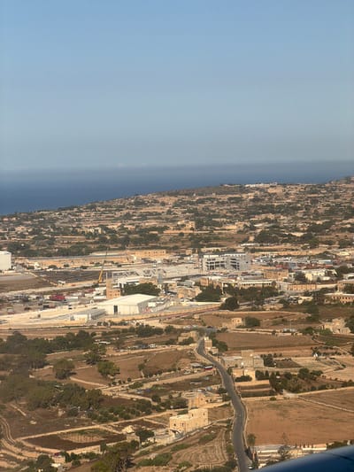 An aerial view of Birżebbuġa, Malta, captured from an elevated perspective. The scene showcases a mix of industrial and rural landscapes. In the foreground, several large, modern buildings with white roofs and metallic structures are visible, indicative of an industrial area. Adjacent to these are fields, some appearing cultivated with brown soil and others with scattered greenery and trees, suggesting agricultural activity. A winding road snakes through the landscape, connecting various buildings and fields.
Further into the mid-ground, a cluster of older, light-colored stone buildings with visible roofs are scattered across a hillside dotted with trees. This area appears to be a more established part of the town or a rural settlement.
The background features a gently sloping hillside densely populated with more of these stone structures and trees, blending into the natural terrain. Beyond the land, a vast expanse of deep blue water, likely the Mediterranean Sea, meets the horizon under a clear, light blue sky. A distant, hazy shoreline or island is faintly visible where the sea meets the sky. The overall impression is a coastal town with a significant industrial presence integrated with a more traditional, rural environment.