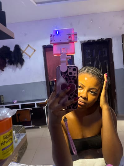 A young adult female with dark skin and dark braided hair (cornrows) is depicted in a mirror selfie, taken indoors in Gbongan, Nigeria. She wears a dark, strapless top. Her right hand, with red manicured nails, holds a smartphone in a pink and black leopard-print case, which is attached to a device emitting pink and blue light, likely a selfie stick with an integrated ring light. A pink wrist strap hangs from the phone. Her left hand is positioned near her left ear. She maintains a neutral expression, looking directly towards the camera. The reflected background reveals a domestic interior with light-colored walls, a dark paneled door, and a light-colored tiled floor. On the left, dark hair extensions or wigs hang from a wooden rack on the wall, above a dark cabinet with lighter panels. An orange container, possibly a cosmetic product, with partially visible text "Nodra," is reflected in the lower left foreground.