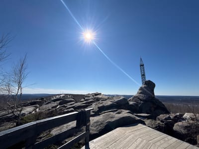 The image shows a sunny, daytime scene from a high vantage point in Gubakha, Russia. Dominating the foreground are large, grey rock formations. A wooden deck with a railing is visible, suggesting a constructed viewing platform. A dark grey garment hangs over the railing. A tall metal structure stands atop one of the rocks, possibly a communications tower or observation point. Beyond the rocks, a landscape of forested hills stretches into the distance, with patches of snow still visible. The sky is clear and bright blue, with the sun shining intensely. Bare branches of trees are visible in the lower left.