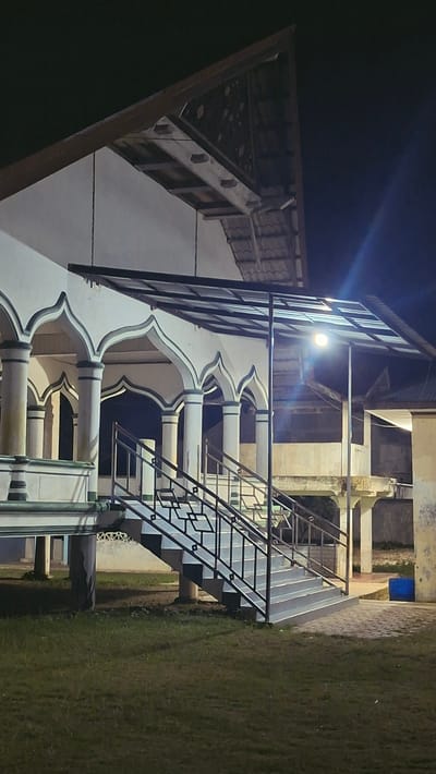A vertical, low-angle, night-time shot captures the exterior of a building with Islamic architectural elements in Keudebayu, Indonesia. The building features a white facade with green trim, accented by a series of arched colonnades. A metal staircase with black railings leads up to an open-air walkway covered by a translucent canopy. A bright floodlight illuminates the area around the stairs. The surrounding environment consists of a dark lawn with patches of bare earth. In the background, a blue plastic bin is visible. The sky is dark, indicating it is night. The scene appears to be a public space, possibly a place of worship or a community building.