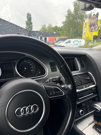 The image shows the interior of an Audi car, viewed from the driver's seat. The steering wheel, with the Audi logo, is prominently displayed. The dashboard features a speedometer and other instruments. A car air freshener shaped like a cat is hanging from the rearview mirror. The windshield is covered in raindrops, suggesting it is raining outside. In the background, through the windshield, a building and trees are visible, along with parked vehicles, including a white van. The location is the city of Vellmar, Germany.