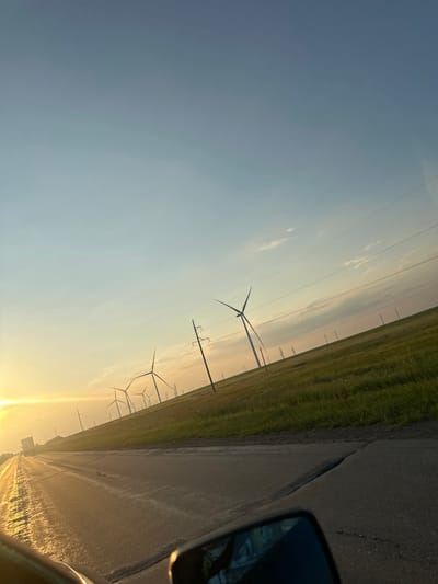 This media file depicts a view from a vehicle driving down a road in a rural, open landscape, likely during either sunrise or sunset. The scene is dominated by a field of wind turbines stretching into the distance, with power lines also visible crisscrossing the sky. The road surface appears wet, reflecting the warm, golden light of the sun. Tall grass lines the side of the road, adjacent to the field where the wind turbines are situated. The sky is a gradient of soft blues and yellows, with some wispy clouds. The overall impression is one of natural energy generation in a vast, open space. The camera angle is tilted, giving a dynamic, sweeping perspective of the scene. There is no visible text in the image.