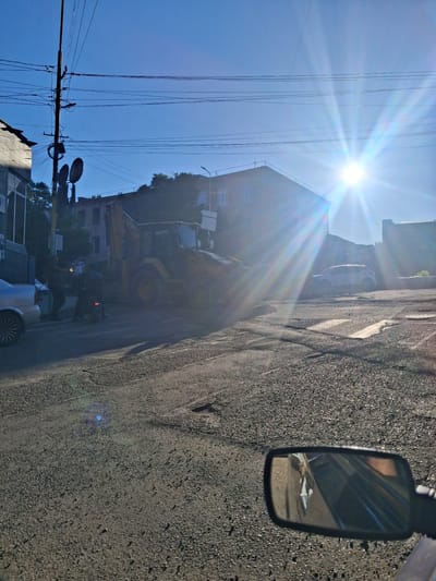 The image shows a street scene in Yeghegnadzor, Armenia, on a bright, sunny day, likely late morning or early afternoon given the sun's position and intensity. The perspective is from inside a vehicle, as evidenced by the side mirror reflecting a portion of the interior and the road ahead.
The foreground is dominated by the asphalt road, showing some wear and tear with cracks and patches. To the left, a clear blue sky stretches out, with a brilliant sun emitting strong rays, creating lens flare across the image. A complex network of power lines crisscrosses the sky, suspended from poles.
In the mid-ground, on the left side of the street, a building with multiple stories is visible, set against a slightly elevated slope. A construction site is in progress, with a yellow backhoe loader and an excavator. Several individuals, appearing as small figures in the distance, are present at the construction site. Further back, other buildings and trees can be seen. A white car is parked or moving on the street.
In the side mirror, a reflection shows part of the vehicle's interior, possibly a dashboard or seating.
The overall impression is a typical urban or semi-urban scene in Armenia, with active construction work taking place under a clear, sunny sky. The strong sunlight suggests good weather. No specific text is clearly legible in the image, though the backhoe loader might have some markings that are too small to read.