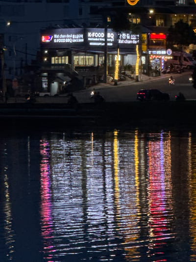 The image captures a nighttime view of a waterfront area in Xuân Hương Lake, Đà Lạt, Vietnam. The foreground is dominated by the dark water of the lake, reflecting the vibrant lights of buildings and streetlights along the shore. These reflections create abstract patterns of red, yellow, blue, and white light shimmering on the water's surface.
In the background, a row of buildings is illuminated by neon signs and interior lighting. One prominent sign reads "K-FOOD BBQ SINCE 2008" in English and Korean characters. Other signs are partially visible, suggesting a commercial area with restaurants and shops. Two trees adorned with yellow string lights stand on the sidewalk in front of the buildings, adding to the festive atmosphere.
A dark-colored car is parked on the side of the road in front of the waterfront. Several motorcycles are also present, some parked and one possibly in motion on the street. The overall scene depicts a lively urban environment at night, with a focus on the interplay between artificial lights and the natural element of the lake.